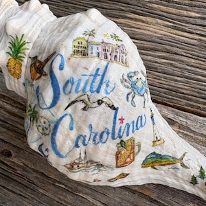 May include: A large, white conch shell decorated with colorful illustrations and the words "South Carolina" in blue script. The shell features images of a pineapple, crab, lighthouse, and other coastal elements.