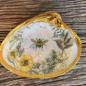 May include: A gold-rimmed, white ceramic dish with a pressed floral design and a bee. The dish is filled with dried flowers and leaves.