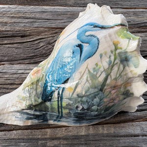 May include: A large, off-white conch shell featuring a detailed, colorful illustration of a blue heron standing among green foliage and small yellow and pink flowers. The shell has a smooth, glossy finish and a natural, irregular, fluted edge.