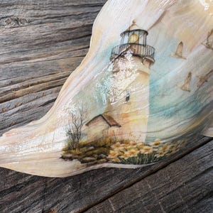 May include: A large, cream-colored conch shell featuring a detailed decal of a lighthouse scene. The illustration depicts a tall lighthouse with a glowing lantern room, surrounded by yellow flowers, bare trees, and several birds in flight.
