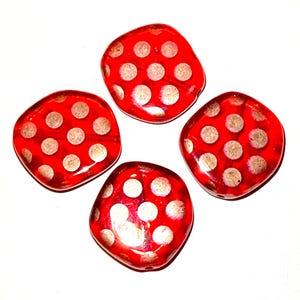May include: Four red, square-shaped glass beads with white polka dots. The beads are arranged in a square pattern against a white background. The beads have a glossy finish and are likely used for jewelry making or crafts.