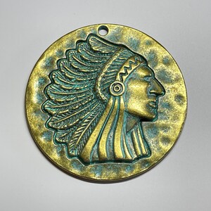 May include: A round, antique-style pendant with a Native American chief profile. The pendant is bronze-colored with a turquoise patina, featuring a textured surface and a small hole at the top for hanging.