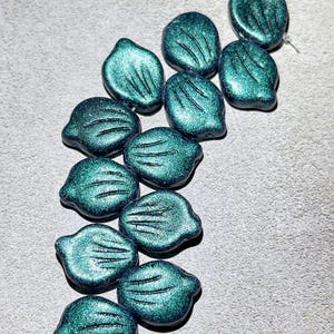 May include: A collection of teal green glass beads with a textured surface. The beads are shaped like leaves and have a metallic sheen.