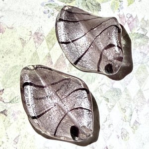 May include: Two clear, leaf-shaped glass beads with black vein-like lines. The beads have a light purple hue and a small hole at the bottom. The background is a floral and diamond pattern.