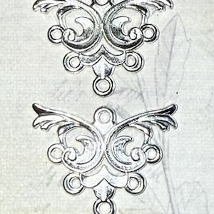 May include: Two silver-colored filigree connectors with an ornate, symmetrical design. Each connector features a central floral element, swirling details, and loops for attachment. The connectors have a wing-like shape.