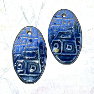 May include: Two blue ceramic oval pendants with gold accents. Each pendant has a geometric pattern and a small hole at the top for hanging. The pendants are likely for jewelry making or crafting.