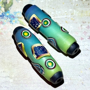 May include: Two handcrafted glass beads with a gradient of blue and green. Each bead features decorative elements including a square with a starry night design and circular accents with yellow and green details. The ends are capped with black.