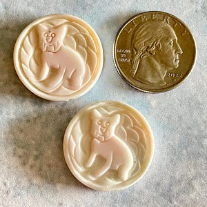 May include: Two white, round, ceramic buttons with a raised image of a cartoon dog. The dog is sitting with its tongue sticking out and has a collar. The buttons are about the size of a quarter.