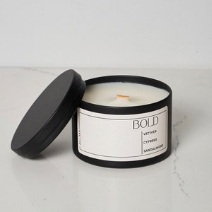 May include: A black candle tin with a white label that reads "BOLD" with the scents "VETIVER, CYPRESS, SANDALWOOD." The lid is off to the side. The candle has a wooden wick.
