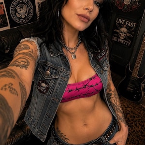 Könnte beinhalten: Eine ärmellose blaue Jeansweste mit Totenkopf-Aufnäher, getragen über einem leuchtend pinken Bandeau-Top mit schwarzem Stacheldraht-Muster. Das Outfit umfasst eine blaue Jeans mit einem nietenbesetzten schwarzen Gürtel und eine silberne Kette mit Vorhängeschloss.