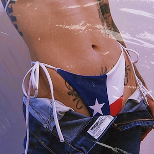 Puede incluir: Una mujer lleva puesto un bikini de color azul, blanco y rojo con un cordón blanco. La parte inferior del bikini tiene una estrella blanca en la sección azul. La mujer lleva unos vaqueros azules sobre la parte inferior del bikini. Los vaqueros tienen una etiqueta de Wrangler.