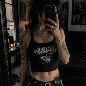 Op de afbeelding: Een zwarte mouwloze crop top met een witte grafische print van een vlieg en de tekst The Cramps Human Fly. Het kledingstuk heeft een ronde hals en wordt gecombineerd met een zwarte broek met een leren riem en metalen kettingen.