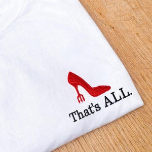 May include: White t-shirt featuring a red high-heeled shoe design, with a fork replacing the heel. The text "That's ALL." is printed in black below the shoe. The shirt is on a wooden surface.