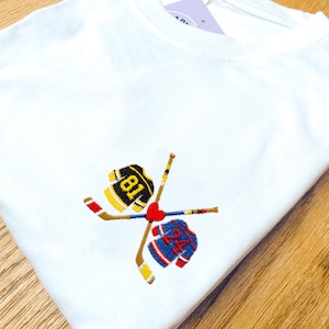 May include: White t-shirt with an embroidered hockey design. The design includes two crossed hockey sticks, two team jerseys with the numbers 81 and 24, and a red heart. The jerseys are black and yellow, and blue and red.