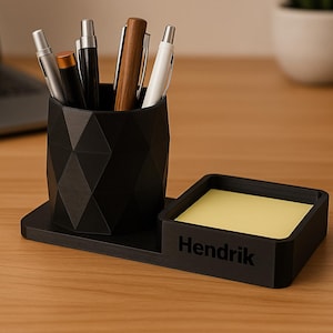 Könnte beinhalten: Schwarzer Schreibtisch-Organizer mit einem geometrischen Stifthalter und einem quadratischen Notizblockhalter. Der Notizblockhalter trägt den Namen "Hendrik". Mehrere Stifte und Bleistifte befinden sich im Stifthalter. Der Organizer steht auf einem Holzschreibtisch.