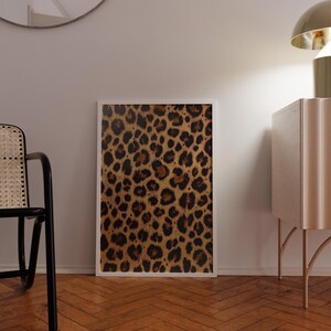 May include: A framed print of a leopard print pattern in shades of brown and black. The print is hanging on a white wall in a room with a wooden floor.