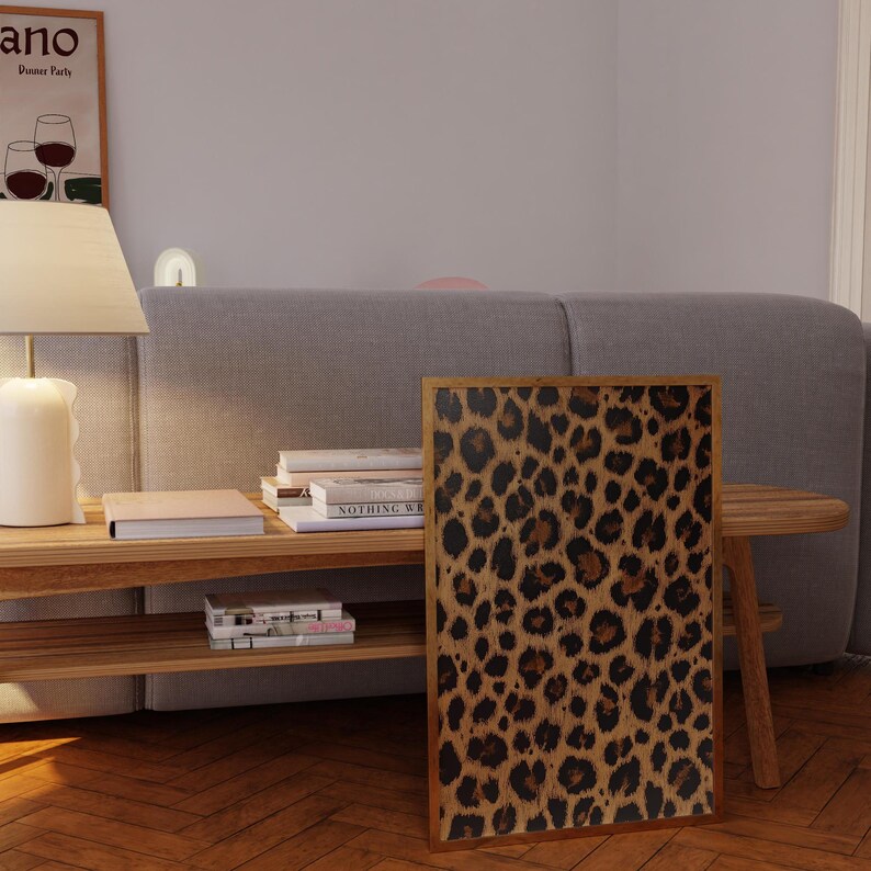 May include: A framed print of a brown and black leopard print pattern. The print is hanging on a wall above a wooden shelf with books and magazines.