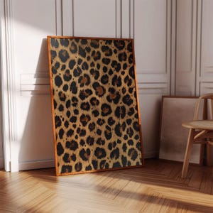 May include: A framed print featuring a leopard print pattern in shades of brown, black, and gold. The print is in a wooden frame and is leaning against a white wall.
