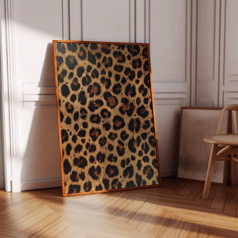 May include: A framed print featuring a leopard print pattern in shades of brown, black, and gold. The print is in a wooden frame and is leaning against a white wall.