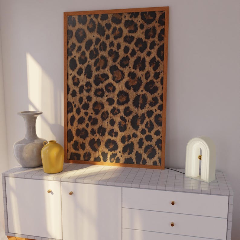 May include: A white dresser with two drawers and a brown framed picture of a leopard print pattern hanging above it. A white ceramic lamp with a gold accent is on the dresser.