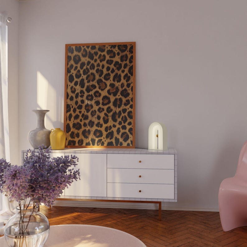 May include: A white ceramic lamp with a gold accent sits on a white dresser with a framed leopard print artwork behind it. A pink chair is in the background.