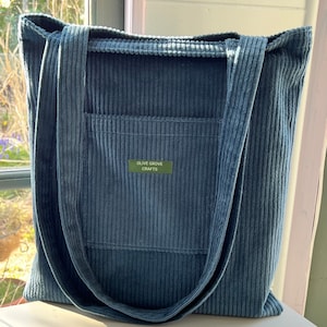 May include: A blue corduroy tote bag with a pocket on the front. The bag has two long straps and a label that reads "Olive Grove Crafts".