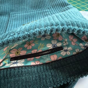 Op de afbeelding: Een tealblauwe corduroy tas met een ritsvak. De tas is gevoerd met een bloemenstof met roze en witte bloemen op een turquoise achtergrond.