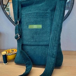 Photograph shows an aqua green corduroy bag with a long shoulder strap resting on a table.  In the background there is a mirror on the wall and a reel of cotton in matching colour is placed to the side of the bag