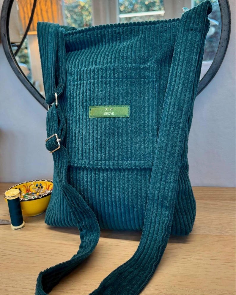Photograph shows an aqua green corduroy bag with a long shoulder strap resting on a table.  In the background there is a mirror on the wall and a reel of cotton in matching colour is placed to the side of the bag