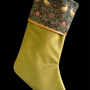 May include: A Christmas stocking featuring a solid olive green velvet body and a contrasting cuff. The cuff displays a dark background with a repeating pattern of light-coloured birds, pink flowers, and green foliage, finished with a thin gold trim.