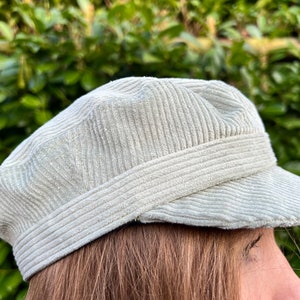 May include: A pale green corduroy cap with a short peak. The hat features a structured, rounded crown and a band detail. The fabric has a ribbed texture. The cap is set against a backdrop of green foliage.