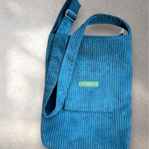 Op de afbeelding: Een blauwe corduroy tote bag met een lange band en een klein vakje aan de voorkant. Het vakje heeft een groen label met de tekst "OLIVE GROVE".