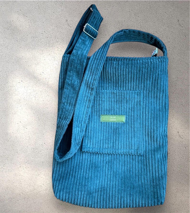 Op de afbeelding: Een blauwe corduroy tote bag met een lange band en een klein vakje aan de voorkant. Het vakje heeft een groen label met de tekst "OLIVE GROVE".