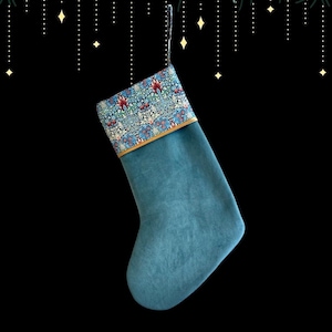 May include: A teal Christmas stocking with a floral patterned cuff. The cuff features a blue, red, and white floral design with a gold trim. The stocking is hanging against a black background with decorative lights.