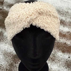 May include: A cream-coloured, textured headband with a twisted design. The headband is made of a soft, curly material and is displayed on a black mannequin head. The background shows snow.