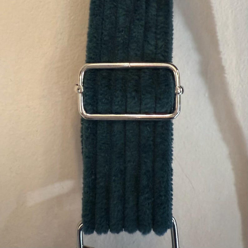 Op de afbeelding: Een donkergroene corduroy riem met een zilveren metalen gesp. De gesp is rechthoekig met afgeronde hoeken.