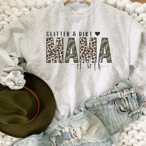 K&ouml;nnte beinhalten: Ein grauer Pullover mit dem Text "Glitter & Dirt Mama of both" in Schwarz und Wei&szlig;. Das Wort "Mama" ist in Leopardenmuster und Tarnmuster.