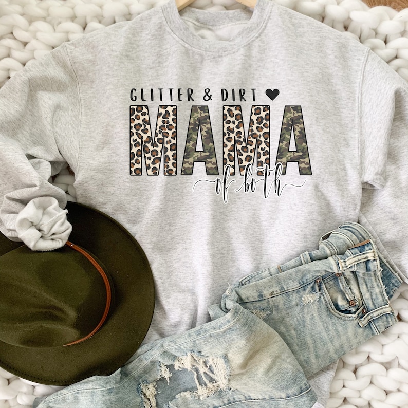 K&ouml;nnte beinhalten: Ein grauer Pullover mit dem Text "Glitter & Dirt Mama of both" in Schwarz und Wei&szlig;. Das Wort "Mama" ist in Leopardenmuster und Tarnmuster.