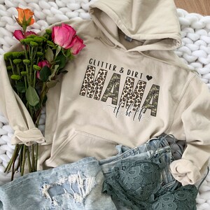 K&ouml;nnte beinhalten: Ein beiger Hoodie mit einem grafischen Aufdruck, der "Glitter & Dirt Mama of Both" in Schwarz, Braun und Gr&uuml;n lautet. Die Buchstaben sind mit Leopardenmuster und Tarnmuster verziert.