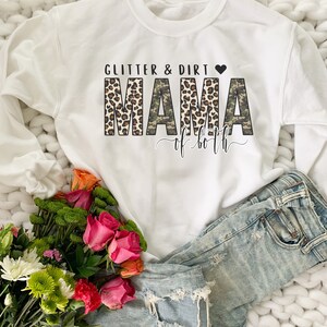 K&ouml;nnte beinhalten: Wei&szlig;er Pullover mit dem Text "Glitter & Dirt Mama of Both" in braunem, schwarzem und wei&szlig;em Leopardenmuster und Tarnmuster. Der Text ist von einem schwarzen Herzen umgeben.