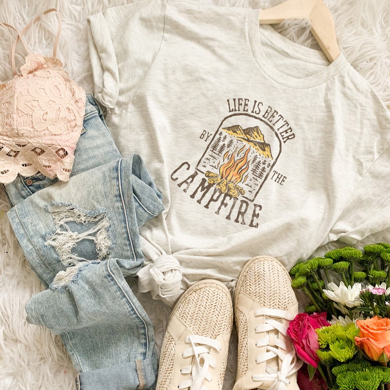 May include: A light gray t-shirt with a graphic of a campfire and the text "Life is better by the campfire". The shirt is laid out on a white fuzzy surface with a pair of white canvas shoes, blue jeans, a pink lace bra, and a bouquet of flowers.