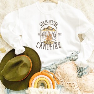 May include: White sweatshirt with the text "LIFE IS BETTER BY THE CAMPFIRE" in a brown graphic. The graphic includes a campfire, mountains, and trees. An olive green hat, a rainbow macrame, and a beige lace bra are also visible.