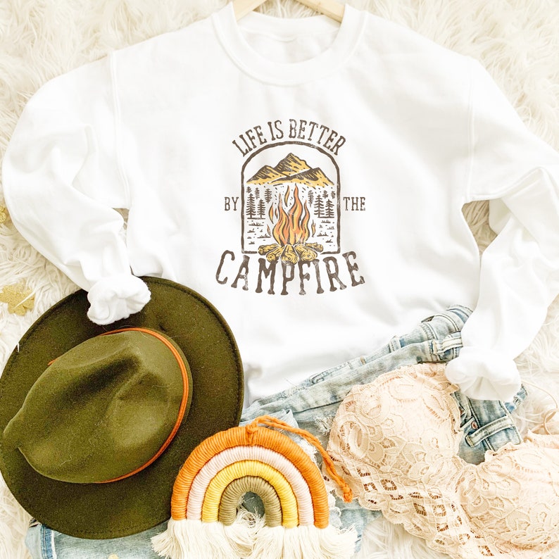 May include: White sweatshirt with the text "LIFE IS BETTER BY THE CAMPFIRE" in a brown graphic. The graphic includes a campfire, mountains, and trees. An olive green hat, a rainbow macrame, and a beige lace bra are also visible.