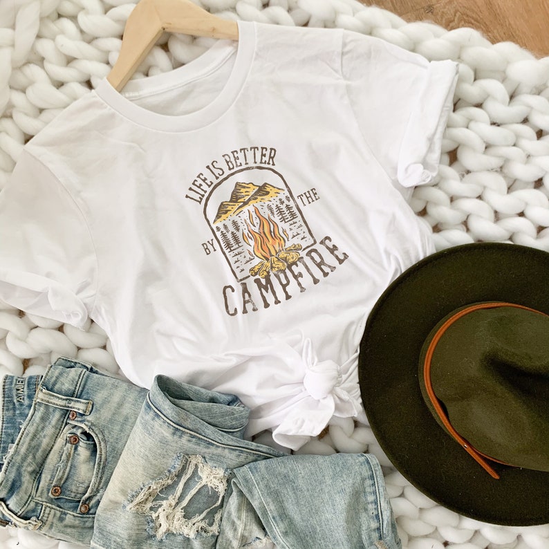 May include: White t-shirt with a graphic design of a campfire in a mountain scene. The text "Life is better by the campfire" is printed on the shirt.