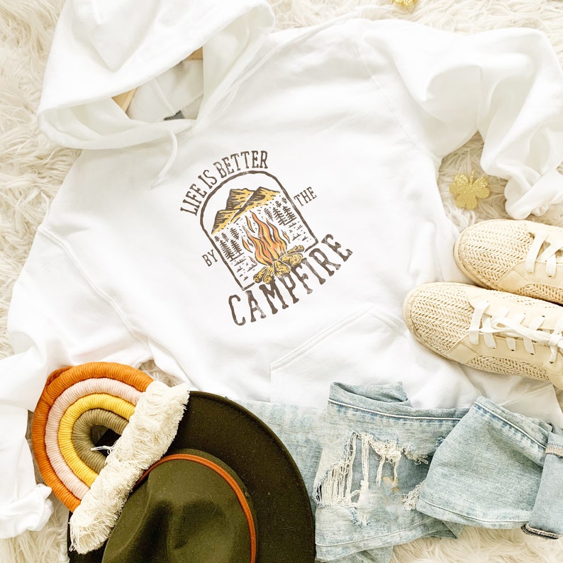 May include: White hooded sweatshirt with the text "LIFE IS BETTER BY THE CAMPFIRE" in brown. The graphic includes a campfire and mountain scene. The image also shows a rainbow wall hanging, a green hat, denim shorts, and beige shoes.