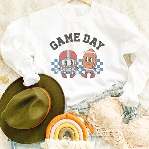 May include: White sweatshirt with a graphic of a football helmet and football with the text "GAME DAY" in black letters.
