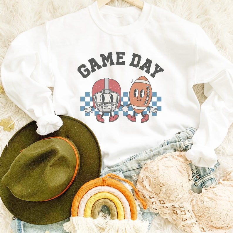 May include: White sweatshirt with a graphic of a football helmet and football with the text "GAME DAY" in black letters.