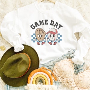 May include: White long-sleeved sweatshirt with the text "GAME DAY" in black lettering. A cartoon baseball and baseball glove are holding hands on a checkered background.