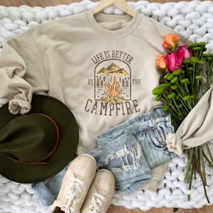 May include: A beige sweatshirt with the text "Life is Better by the Campfire" in brown. The graphic features a campfire, mountains, and trees. The sweatshirt is paired with light-wash distressed jeans, beige shoes, and a green hat. Flowers are also present.
