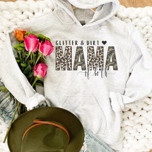 K&ouml;nnte beinhalten: Ein grauer Hoodie mit einer schwarzen und wei&szlig;en Grafik, auf der "Glitter & Dirt Mama of both" mit einem Leopardenmuster und einem Tarnmuster steht.